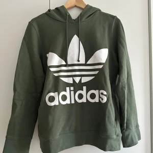 Grön hoodie från Adidas - Säljer en snygg grön hoodie från Adidas med det klassiska vita logotyptrycket på framsidan. Den har en bekväm passform med långa ärmar och en praktisk huva med dragsko samt fickor framtill. Perfekt för både vardag och träning