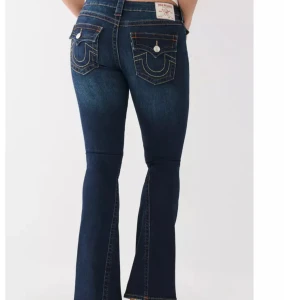 Mörkblå jeans från True Religion - Säljer ett par snygga mörkblå jeans från True Religion i modellen Joey Low Rise Flare. De har en låg midja och utsvängda ben, perfekt för en avslappnad stil. Jeansen har coola detaljer med kontrastsömmar och märkeslogga på bakfickorna. Passar perfekt för både vardag och fest!