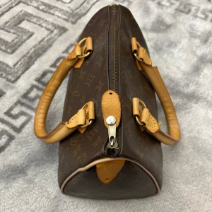 Brun handväska med monogram - Säljer en snygg brun handväska med klassiskt monogrammönster. Väskan har ljusbruna handtag och detaljer, samt en dragkedja upptill. Perfekt för både vardag och fest!