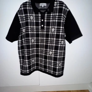 Polo tröja Heaven Can Wait  - Svartvit rutig polotröja med dödskallar från Heaven Can Wait . Tröjan har en klassisk polokrage och knappar i fram. Den är perfekt för att skapa en cool och trendig look.  Bra skick, Använd några gånger.  Köparen står för frakt