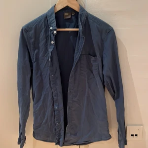 Blå skjorta från ASOS - Säljer en stilren blå skjorta från ASOS i storlek M. Skjortan har långa ärmar och knappar längs hela framsidan. Perfekt för både vardag och festliga tillfällen. Materialet känns mjukt och bekvämt, vilket gör den idealisk för höst och vår.