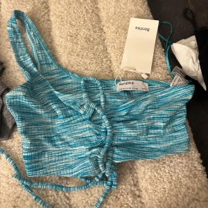 Blå oneshoulder topp från Bershka - Snygg blå oneshoulder topp från Bershka i ett lätt och luftigt material. Toppen har en cool snörning framtill som ger en unik look. Perfekt för sommarens alla fester eller en dag på stan. Helt ny med tag kvar!