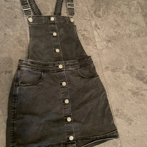 Svart jeansklänning med knappar - Snygg svart jeansklänning med knappar framtill och justerbara axelband. Perfekt för en avslappnad stil, kan matchas med en t-shirt under. Klänningen har en tight passform och är i mycket bra skick. Den är i storlek 158 men passar en XS