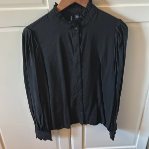 Svart blus med krås - Svart blus från Vero Moda med krås i hals och armslut. Glansigt tyg. Mycket sparsamt använd. 