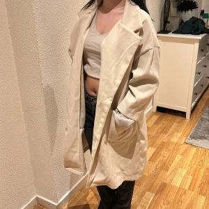 Beige kappa - Säljer en stilren beige kappa i normal passform. Den har långa ärmar och knappar framtill. Perfekt för höst och vår, och ger en elegant touch till vilken outfit som helst.
