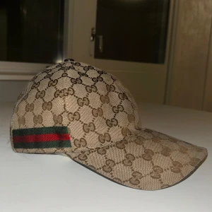 Beige keps från Gucci - Snygg beige keps från Gucci med det klassiska GG-mönstret. Kepsen har en grön och röd randdetalj på sidan. Perfekt för att ge din outfit en lyxig touch. Är äkta! Använt men i mycket bra skick.