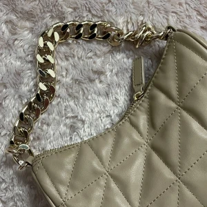 Beige quiltad axelväska med kedja - Snygg beige axelväska i quiltat mönster från Glitter, med en elegant guldfärgad kedja som axelrem. Väskan har en dragkedja upptill för säker förvaring. Perfekt för att ge din outfit en stilren touch.