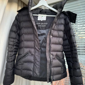 Svart dunjacka från Boomerang  - Snygg svart dunjacka från New Grindo med dragkedja och knappar. Jackan har en quiltad design och avtagbar huva. Perfekt för kyliga dagar.