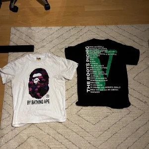 Vit och svart t-shirt från A Bathing Ape och Roots Picnic - Två coola t-shirts till salu! Den vita t-shirten från A Bathing Ape har ett färgglatt kamouflagemönster med deras ikoniska logga. Den svarta t-shirten har texten 'Roots Picnic' i grönt och en lista med artister på baksidan. Perfekta för en avslappnad stil. 300kr: st