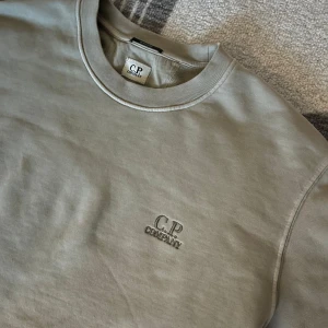Beige tröja från C.P. Company - Säljer en stilren beige tröja från C.P. Company. Tröjan har rund hals och långa ärmar, perfekt för en avslappnad look. Märkets logga är diskret placerad på bröstet. Passar bra till både jeans och chinos. Helt sjukt grisch.