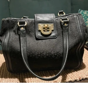 Svart handväska från DKNY - Snygg svart handväska från DKNY med monogrammönster och guldfärgade detaljer. Väskan har en praktisk stängning med ett metallspänne och dubbla handtag för enkel bärning. Perfekt för att ge en elegant touch till din outfit. Kan tänka mig sälja för 250 vid snabb affär