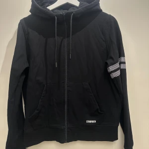 Svart hoodie från Stronger - Säljer en svart hoodie från Stronger med dragkedja och huva. Den har en vit randdetalj på armen och fickor framtill. Perfekt för en avslappnad stil.