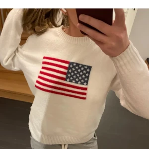 Vit tröja med amerikansk flagga - Säljer en vit tröja med en stor amerikansk flagga på bröstet. Tröjan har långa ärmar och är gjord i ett mjukt material. Perfekt för en avslappnad stil.