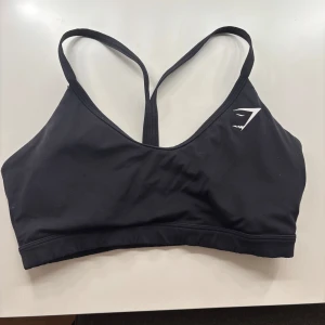 Svart sporttopp från Gymshark - Snygg och bekväm svart sporttopp från Gymshark med korsade axelband och en liten logga på framsidan. Perfekt för träning och ger bra stöd.