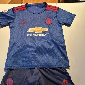 Blå Manchester United fotbollströja och shorts - Säljer en blå Manchester United fotbollströja och shorts med röda detaljer och Chevrolet-logga på framsidan. Tröjan har korta ärmar och Ibrahimovic tryckt namn på baksidan.