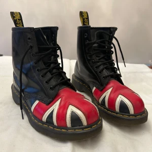 Svarta dr martens boots med Union Jack-mönster - Snygga svarta boots från Dr. Martens med Union Jack-mönster i rött, vitt och blått på tån. Skorna har klassisk snörning och den ikoniska gula sömmen runt sulan. 