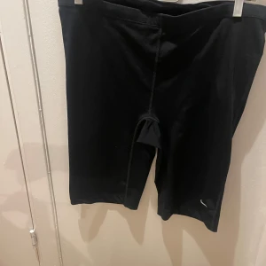 Svarta träningsshorts från Nike - Säljer ett par svarta träningsshorts från Nike. De är knälånga och har en tight passform, perfekta för gymmet eller löprundan. Shortsen har en diskret logga på benet.