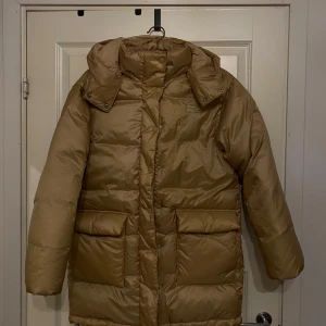 Beige/guld dunjacka med huva - Beige dunjacka med huva. Jackan har långa ärmar och stora fickor framtill. Supervarm, använd 1 gång. Nypris: 2000kr
