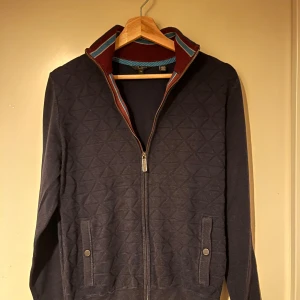 Mörkblå quiltad jacka från Ted Baker - Snygg mörkblå kofta från Ted Baker med dragkedja och ribbad krage i röd och blå randig design. Perfekt för höst och vår med sina långa ärmar och stilrena look.