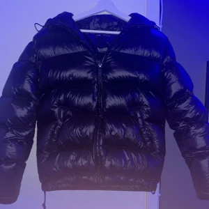 Svart pufferjacka från Zara - Snygg svart pufferjacka från Zara med huva och dragkedja. Perfekt för kyliga dagar med sin vadderade design som håller dig varm. Jackan har en glansig finish och långa ärmar. Det finns ett minihål i ena ärmen som lätt går att laga