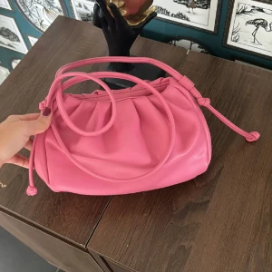 Rosa axelväska i syntet - Snygg rosa axelväska i syntetmaterial med rynkade detaljer och justerbar axelrem. Perfekt för att ge en färgklick till din outfit. (OBS) färgen har lossnat lite inuti men inget man lägger märke till i helhet 