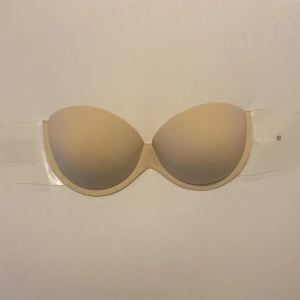 Beige axelbandslös bh med push-up - Helt ny och oanvänd, köpte fel bh därför vill behöver jag sälja den. Den är axelbandslös,  i storlek D-cup, med självhäftande sidor. Perfekt för klänningar och toppar utan axelband. Enkel att använda och ger bra stöd.