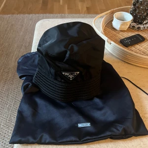 Svart bucket hat från Prada - Snygg svart bucket hat från Prada med deras ikoniska logotyp framtill. Hatten är tillverkad i ett slitstarkt material och har en stilren design.
