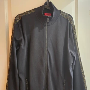 Hugo boss sweatshirt med dragkedja  - Snygg svart sweatshirt med texten hugo boss i guld på ärmarna! Verkar inte säljas längre samt riktigt unik jämfört med de mer basic hugo boss tröjorna! I väldigt fint skick, Använd ett fåtal gånger bara! Nypris närmare 3000kr!
