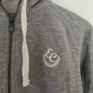 Grå hoodie från Equestrian Stockholm - Säljer en grå hoodie från Equestrian Stockholm med dragkedja och luva. Den har en broderad logga på bröstet och fickor framtill. Snygg och i bra skick!