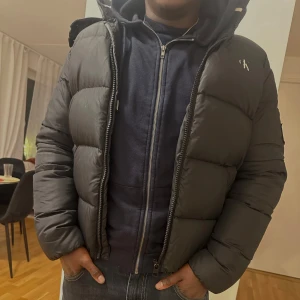 Svart pufferjacka från Calvin Klein - Snygg svart pufferjacka från Calvin Klein med dragkedja och huva. Jackan har märkets logga på bröstet och ryggen. Perfekt för kyliga dagar. Jackan har dock förlorat lite av sitt print på bröstet, men är fortfarande i mycket bra skick