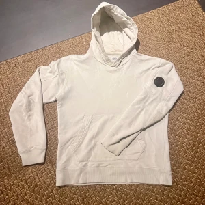 Vit hoodie från C.P. Company - Säljer en stilren hoodie från C.P. Company med en klassisk känguruficka och en rund logodetalj på ärmen. Perfekt för en avslappnad look.