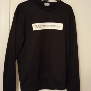 Svart tröja från Emporio Armani - Snygg svart tröja från Emporio Armani med EA7-logga på bröstet och nacken. Perfekt för en stilren look. Tröjan har långa ärmar och en klassisk passform. Storlek XS. PRIS KAN DISKUTERAS!🙏