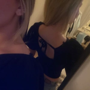 Svart topp med öppen rygg och knyt/rosett - Snygg svart topp med v-ringning och korta ärmar. Toppen har en stilfull öppen rygg med knytband som ger en unik detalj. Perfekt för en kväll ute eller en speciell tillställning.