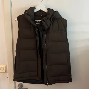 Brun dunväst från Everest - Säljer en brun dunväst från Everest med dragkedja och avtagbar huva. Västen är perfekt för kyligare dagar och har en stilren design med diskret logga på bröstet. Passar utmärkt för höst och vinter.