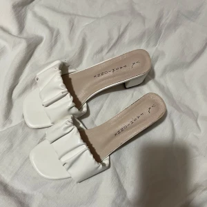 Vita klackskor med volangdetalj - Snygga vita klackskor med en chic volangdetalj över foten. Skorna har en fyrkantig tå och en bekväm blockklack, perfekta för en stilren look.