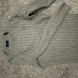 Grå ribbad tröja från GANT - Säljer en stilren grå ribbad tröja från GANT. Tröjan har en liten broderad logga på bröstet och är långärmad med rund halsringning. Perfekt för en avslappnad och snygg look.
