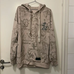 Beige hoodie med tryck - Säljer en beige hoodie med ett unikt tryck. Hoodien har dragkedja och justerbar huva med snören. Perfekt för en avslappnad stil.