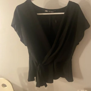 Svart omlottopp från Zara - Snygg svart omlottopp från Zara med knytning i midjan. Toppen har korta ärmar och en elegant v-ringad design. Perfekt för en stilren look.