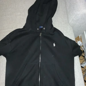 Svart hoodie från Polo Ralph Lauren - Säljer en stilren svart hoodie från Polo Ralph Lauren med dragkedja och klassisk logga på bröstet. Perfekt för en avslappnad look. Den har en praktisk huva och fickor framtill.