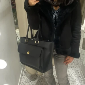 Svart axelväska med gulddetaljer - Snygg svart axelväska i syntetmaterial med guldfärgade detaljer. Väskan har en praktisk dragkedja och ett vridlås framtill. Insidan är rymlig med flera fack för enkel organisering. Får plats med dator o mycket annat. Perfekt för både vardag och speciella tillfällen. Några gropar på framsidan men inget som syns jättemycket i verkligheten