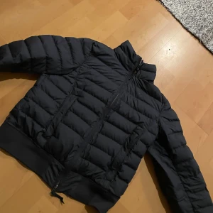 Svart pufferjacka från Parajumpers - Säljer en svart pufferjacka från Parajumpers med dragkedja och broderad logga på ärmen. Jackan har en quiltad design och är perfekt för kyligare väder. Den har långa ärmar och en hög krage för extra värme.