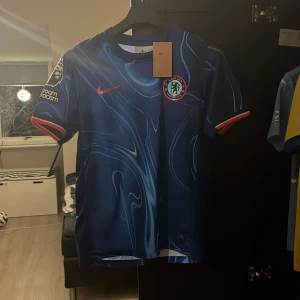 Blå Chelsea tröja från Nike - Snygg blå fotbollströja från Nike med ett unikt mönster och klubbmärke på bröstet. Tröjan har korta ärmar och är tillverkad i Dri-FIT-material för optimal komfort. Perfekt för fotbollsfans som vill visa sitt stöd med stil.