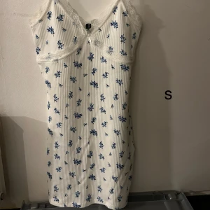 Vit blommig klänning med spetsdetaljer - Charmig vit klänning med blå blommönster och spetsdetaljer vid halsringningen. Klänningen är ärmlös och har en smickrande passform. Perfekt för varma dagar.