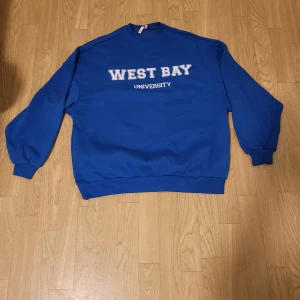 Blå sweatshirt från NLY Trend - Säljer en blå sweatshirt från NLY Trend med texten 'West Bay University' på framsidan. Tröjan har långa ärmar och ribbade muddar vid ärmslut och nederkant. Perfekt för en avslappnad stil. Använd endast ett fåtal gånger