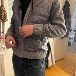 Grå quiltad jacka med stickade ärmar - Säljer en grå quiltad jacka med stickade ärmar och ribbade muddar. Perfekt för kyliga dagar. Okänt märke.