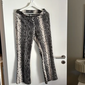 Ormmönstrade byxor i svart och beige - Säljer ett par snygga ormmönstrade byxor i svart och beige. De har en cool och unik design som verkligen sticker ut. Perfekta för en utekväll eller när du vill ha något extra. Byxorna är i storlek S och har en bekväm passform.