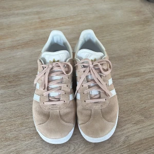 Beige Adidas Gazelle i mocka - Säljer ett par snygga beige Adidas Gazelle sneakers i mocka. De har de klassiska vita ränderna på sidan och 'GAZELLE' i guldtext. Skorna är i bra skick och perfekta för både vardag och fest. De har en vit sula och snörning framtill.