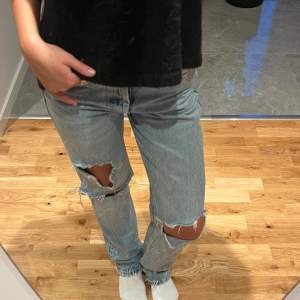 Säljer ett par snygga blå jeans med coola slitningar på knäna. De har en straight passform och är perfekta för en avslappnad stil. Jeansen har en klassisk femficksdesign och knappar i midjan. Perfekta för både vardag och fest!