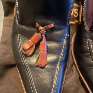 Svarta loafers från Tod's med tofsar - Säljer ett par eleganta svarta loafers från Tod's i skinn. Skorna har dekorativa bruna tofsar på ovansidan och en robust klack. De är i bra skick och perfekta för både vardag och fest.