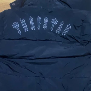 Svart pufferjacka från Trapstar - Säljer en svart pufferjacka från Trapstar i storlek M. Jackan har en cool broderad logga på ryggen och en rund emblem på bröstet. Den är perfekt för kalla dagar med sin quiltade design och dragkedja framtill. Passar både till vardags och fest!